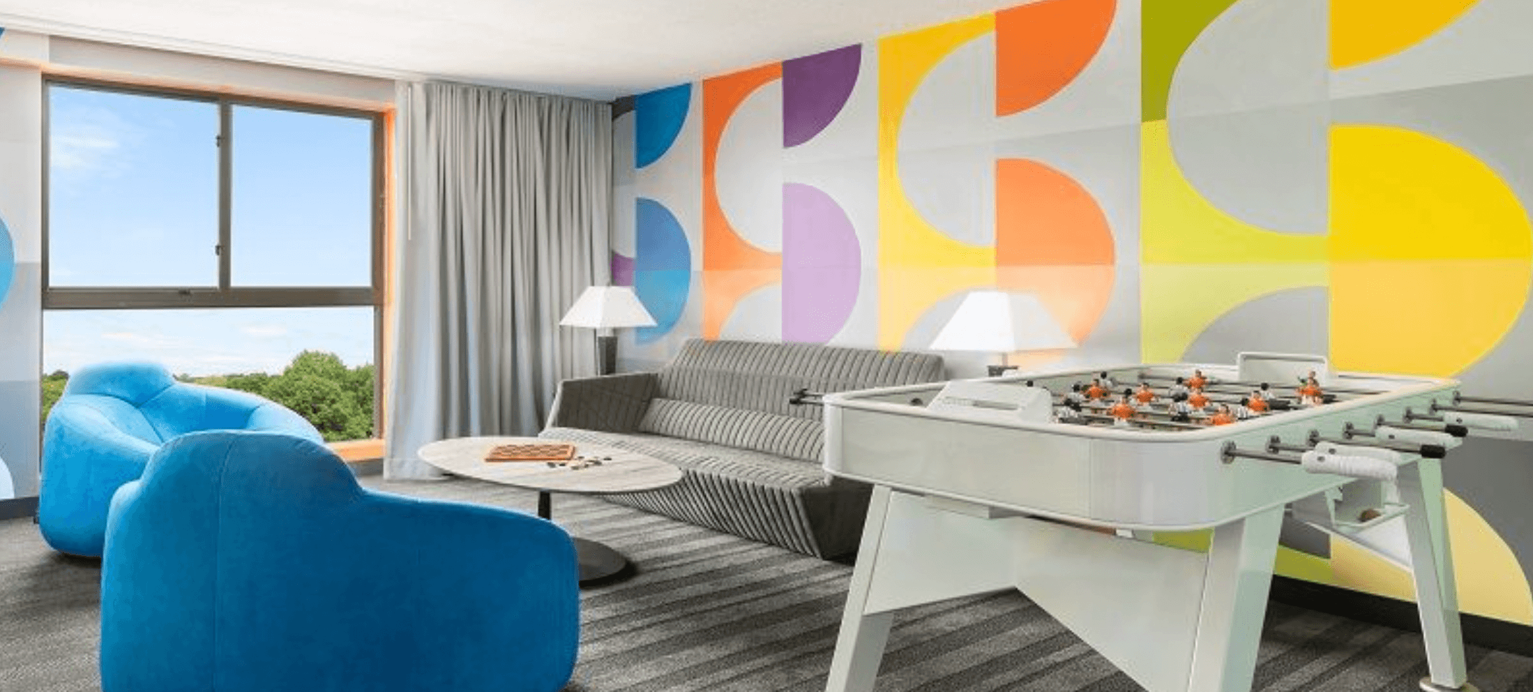 A game room of one of the student-hotels that cogito students stay in for thanksgiving break. It has a grey sofa, two blue armchairs, a table in the middle and a foosball in colorful wall as background.