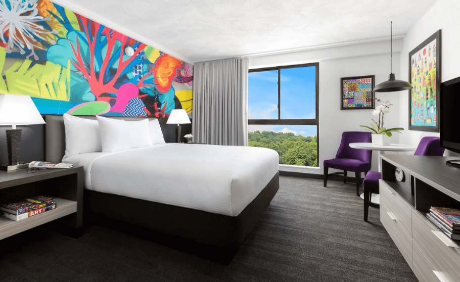 Single hotel room we provide for students in the thanksgiving break program in Boston. A multicolor wall behind the twin bed and a big window in the left. In front of the bed, there is a table with two purple chairs, two paintings and a television.