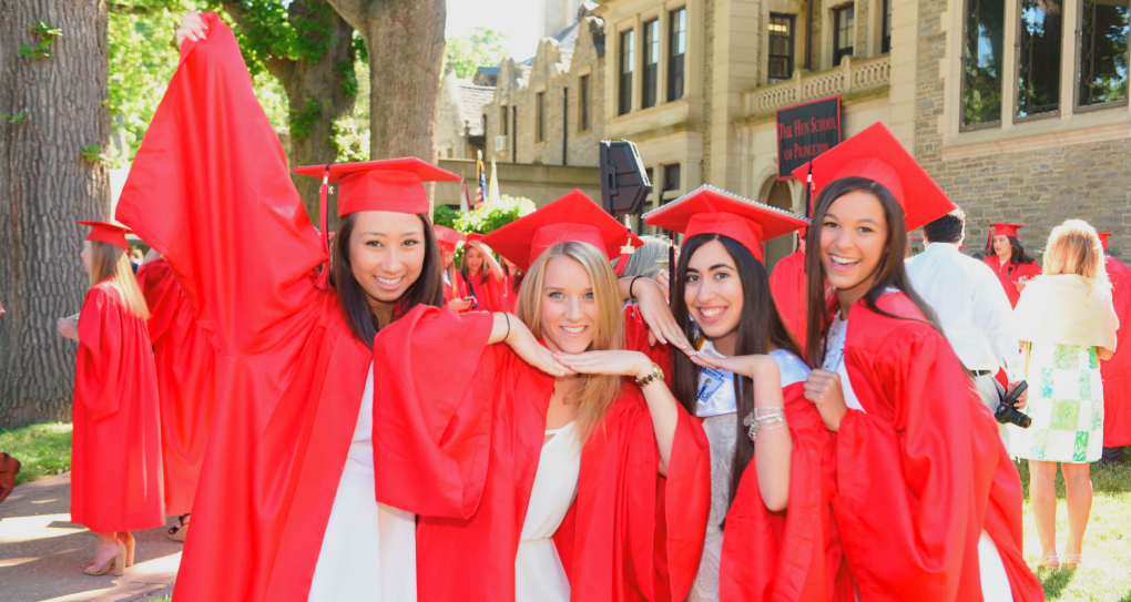 four Cogito female teenagers in red caps and gowns smiling and standing in front of a school building